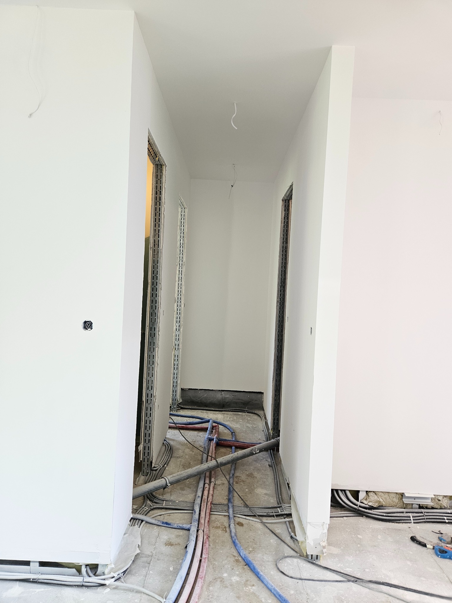 Korytarz w trakcie remontu, widoczne przewody elektryczne i rury instalacyjne na podłodze, ściany pomalowane na biało, ościeżnice drzwiowe w trakcie montażu.
