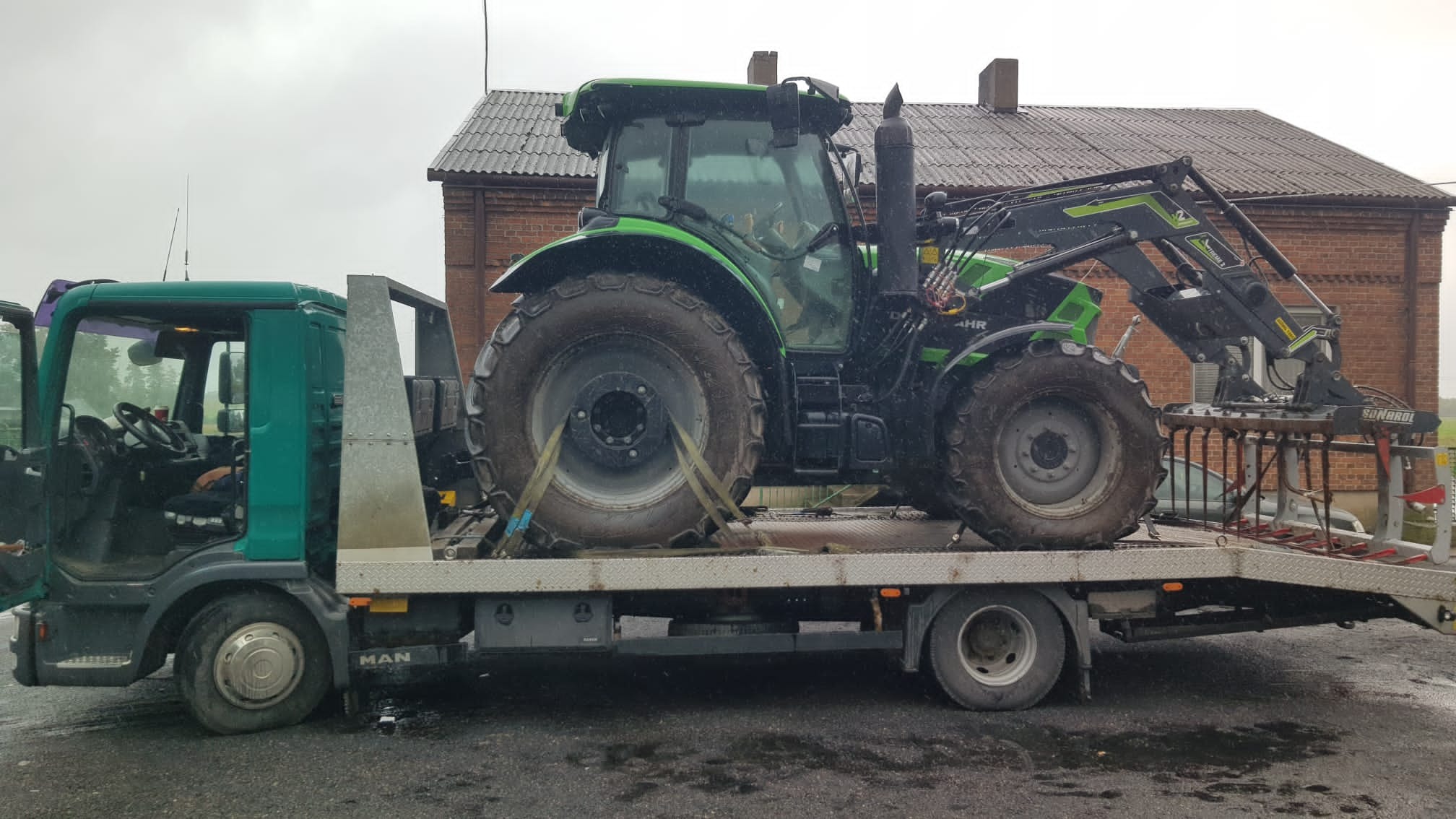 Zielony traktor z zamontowanym z przodu osprzętem rolniczym, zabezpieczony pasami, przewożony na platformie lawety MAN w pochmurny dzień.