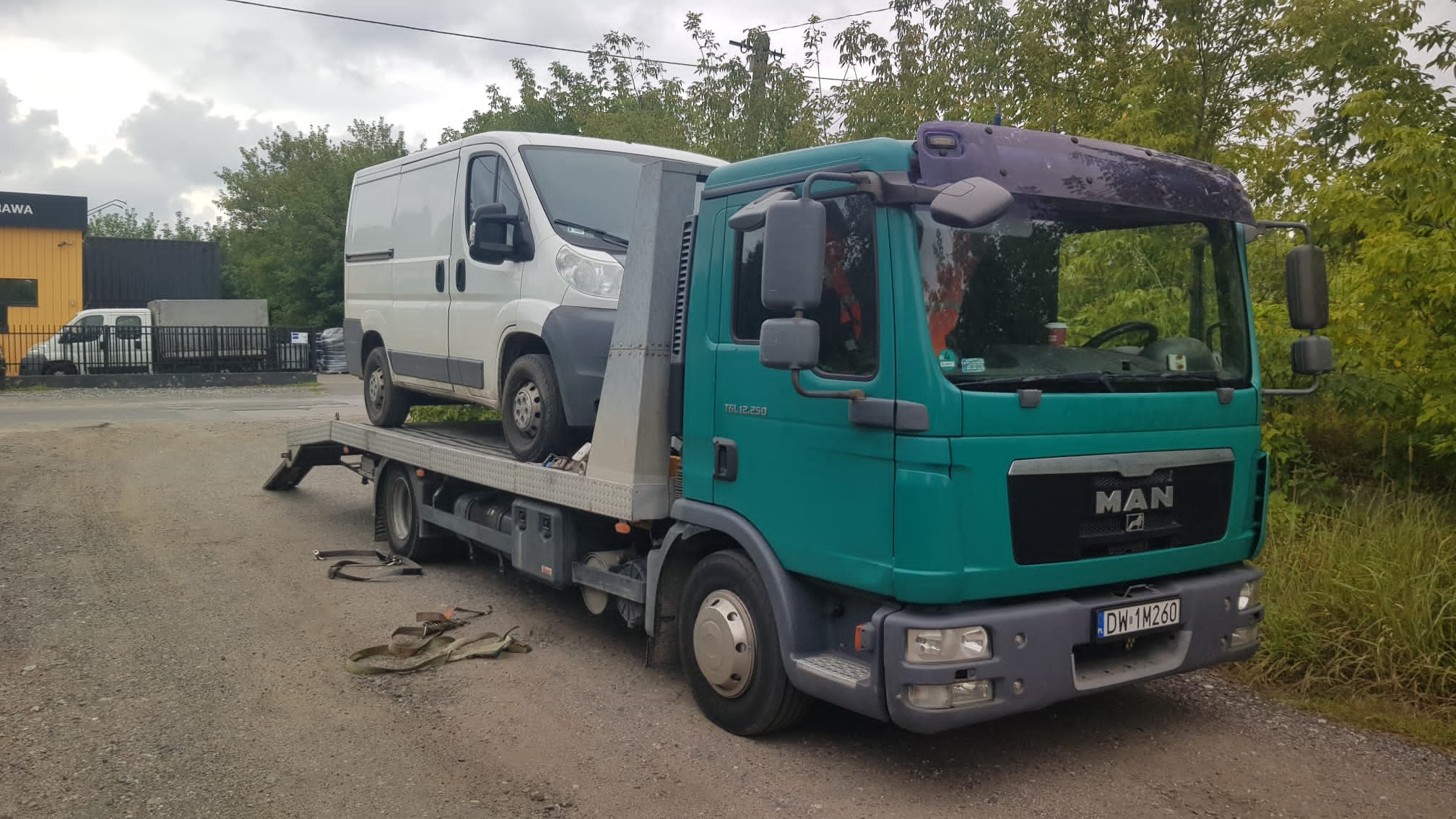 Zielono-szara laweta MAN z białym busem na platformie, zaparkowana na żwirowej drodze obok zieleni, z pasami zabezpieczającymi leżącymi obok.