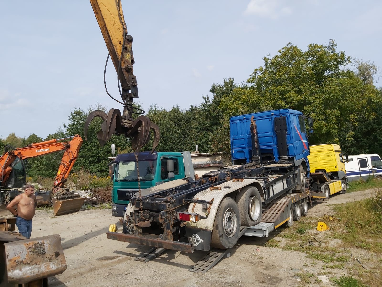Pomarańczowa koparka Hitachi i żuraw z chwytakiem nad zieloną ciężarówką MAN na tle drzew, z niebieską ciężarówką na lawecie za nią i mężczyzną bez koszulki w kadrze.