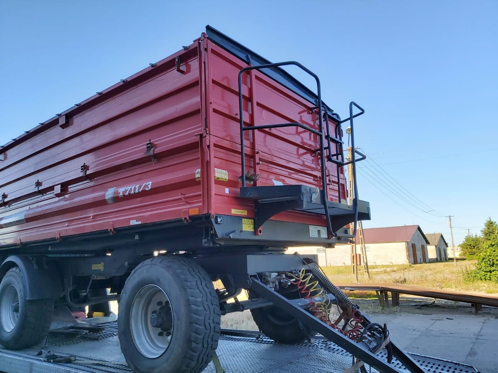 Czerwona przyczepa wywrotka z czarną drabinką z tyłu, zaparkowana na platformie z widocznymi przewodami pneumatycznymi i elektrycznymi, na tle wiejskich budynków i błękitnego nieba.
