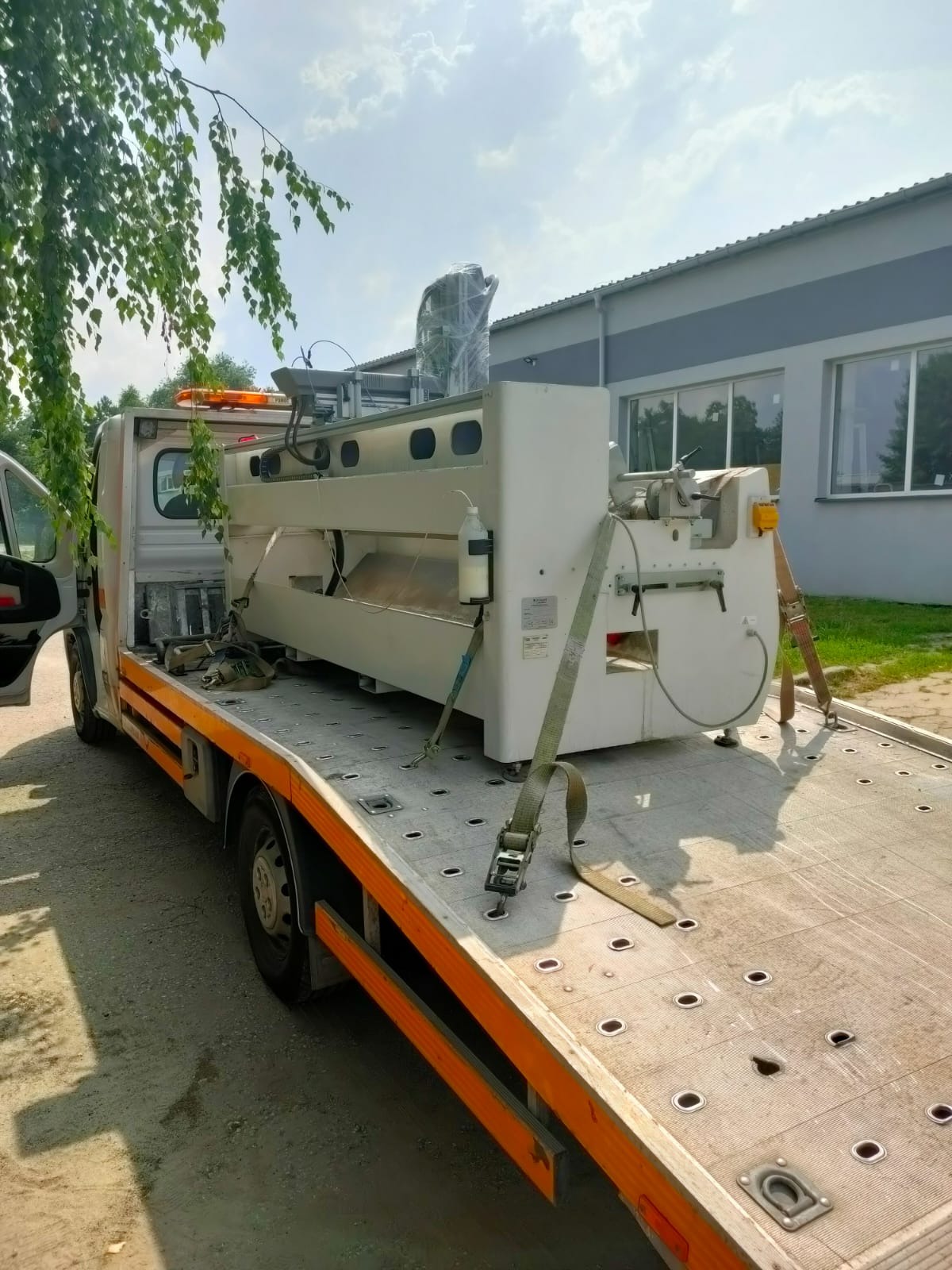 Laweta z zamocowaną maszyną przemysłową, zabezpieczoną pasami transportowymi, na tle budynku przemysłowego.