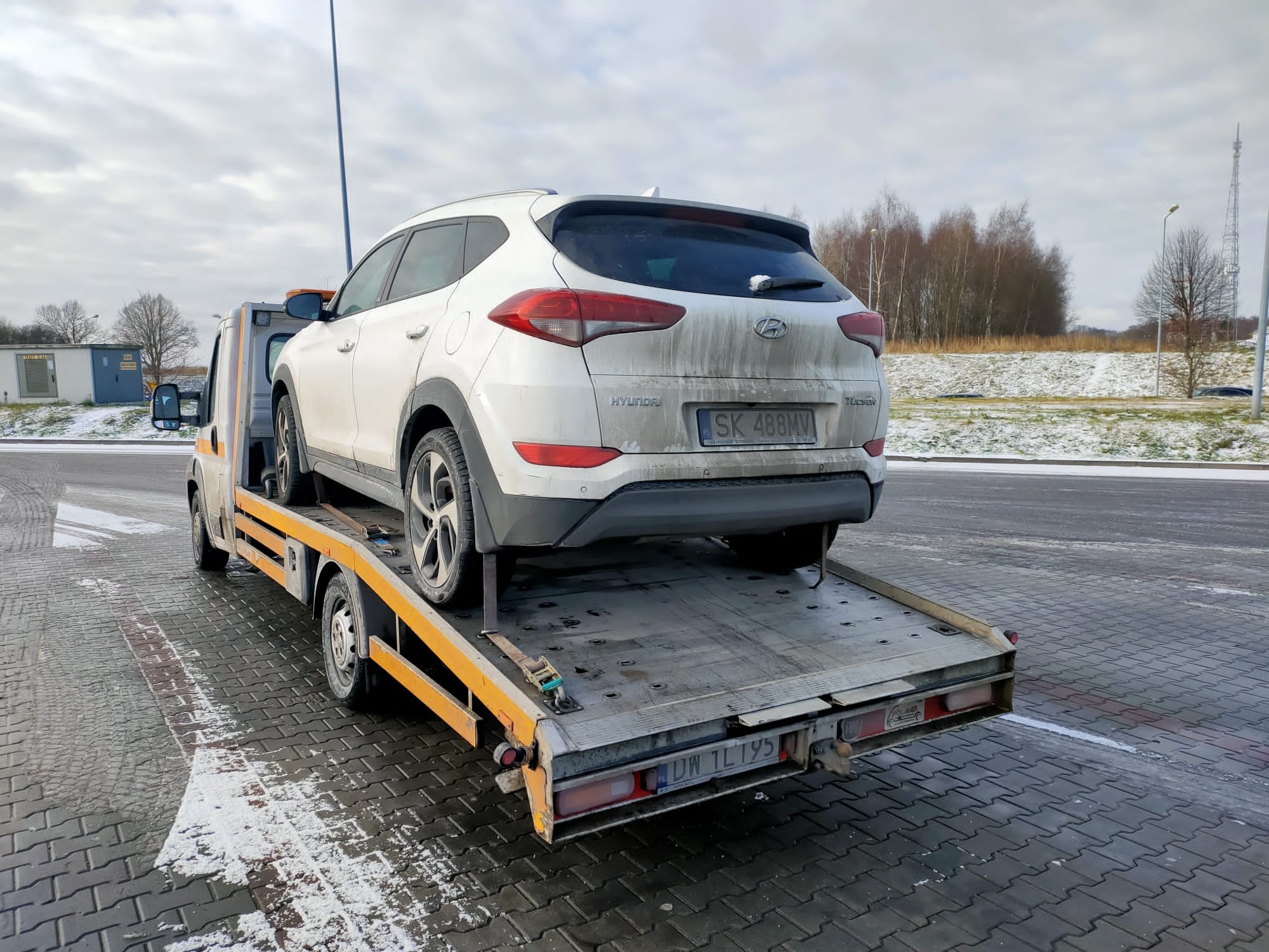 Biały, zabrudzony Hyundai Tucson na platformie lawety, widok z tyłu, tablica rejestracyjna SK 488MV, lekki śnieg na kostce brukowej.