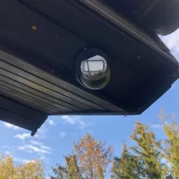 Kamera kopułkowa zamontowana pod okapem dachu, widok z dołu na tle błękitnego nieba i drzew.