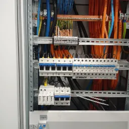 Wnętrze skrzynki elektrycznej z bezpiecznikami Schrack, licznymi przewodami w różnych kolorach (pomarańczowe, niebieskie, czarne, żółto-zielone) i listwami zaciskowymi, zamontowane na szynach DIN.