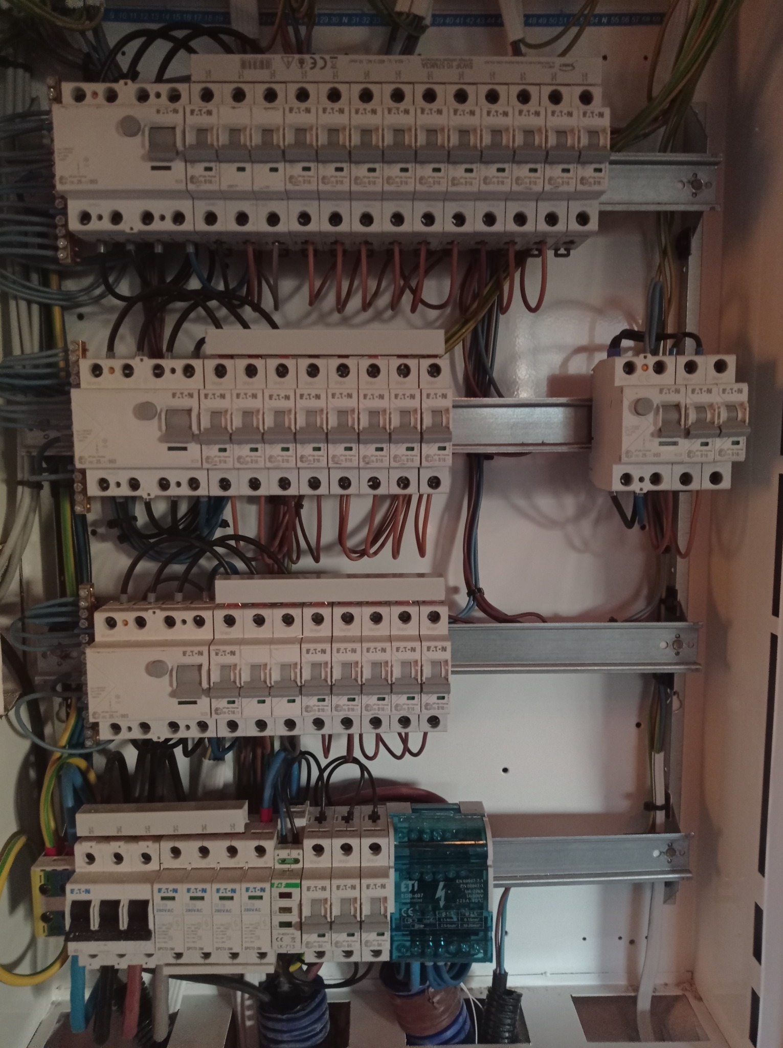 Rozdzielnica elektryczna z widocznymi bezpiecznikami, przewodami i szynami montażowymi, zainstalowana w białej obudowie, z przeważającą ilością przewodów w kolorach czarnym, niebieskim i brązowym.