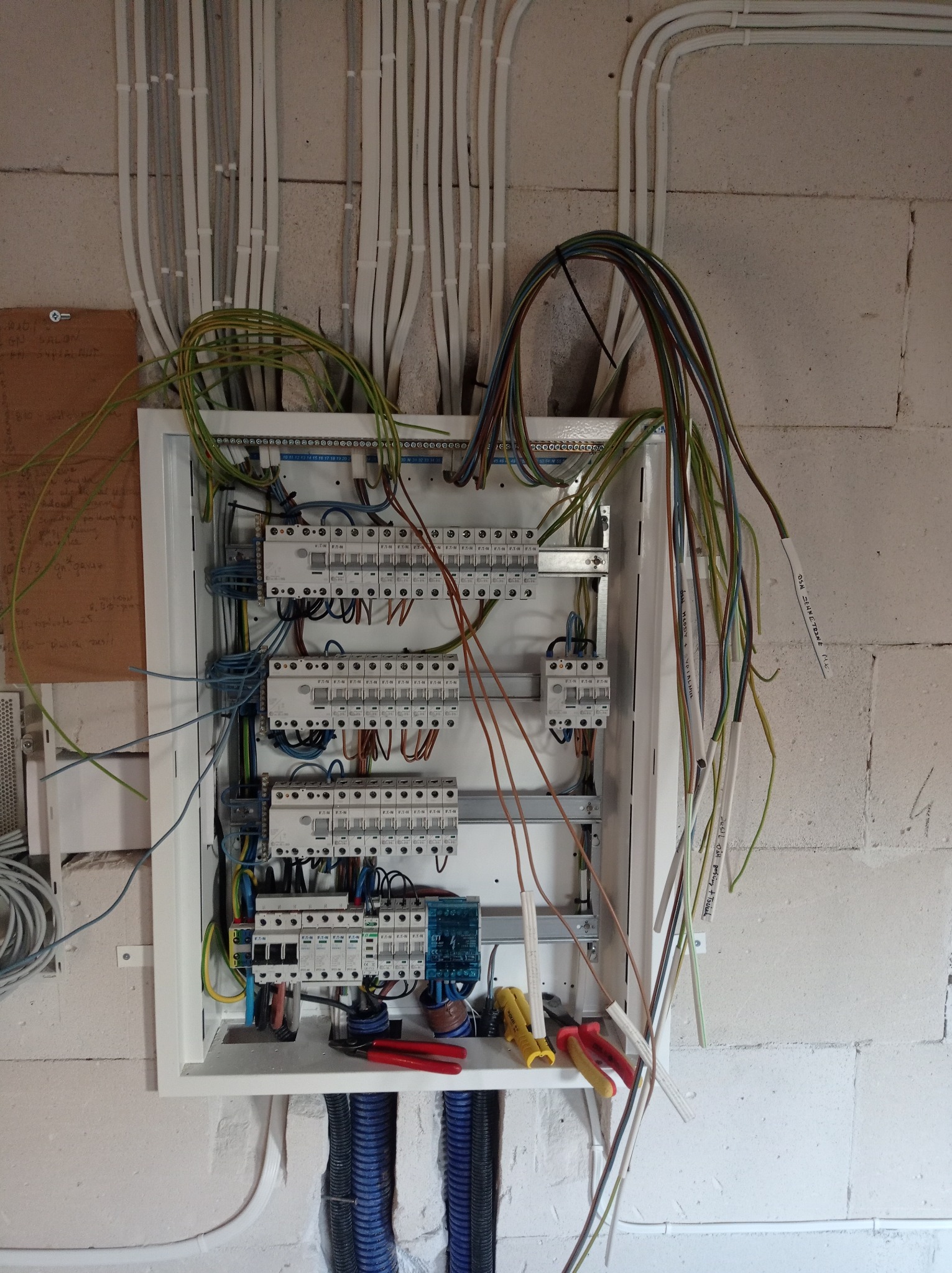Rozdzielnia elektryczna w trakcie montażu, widoczne przewody z opisami, bezpieczniki, narzędzia (kombinerki, obcinaki do kabli) oraz peszle instalacyjne, surowa ściana z betonu komórkowego w tle.