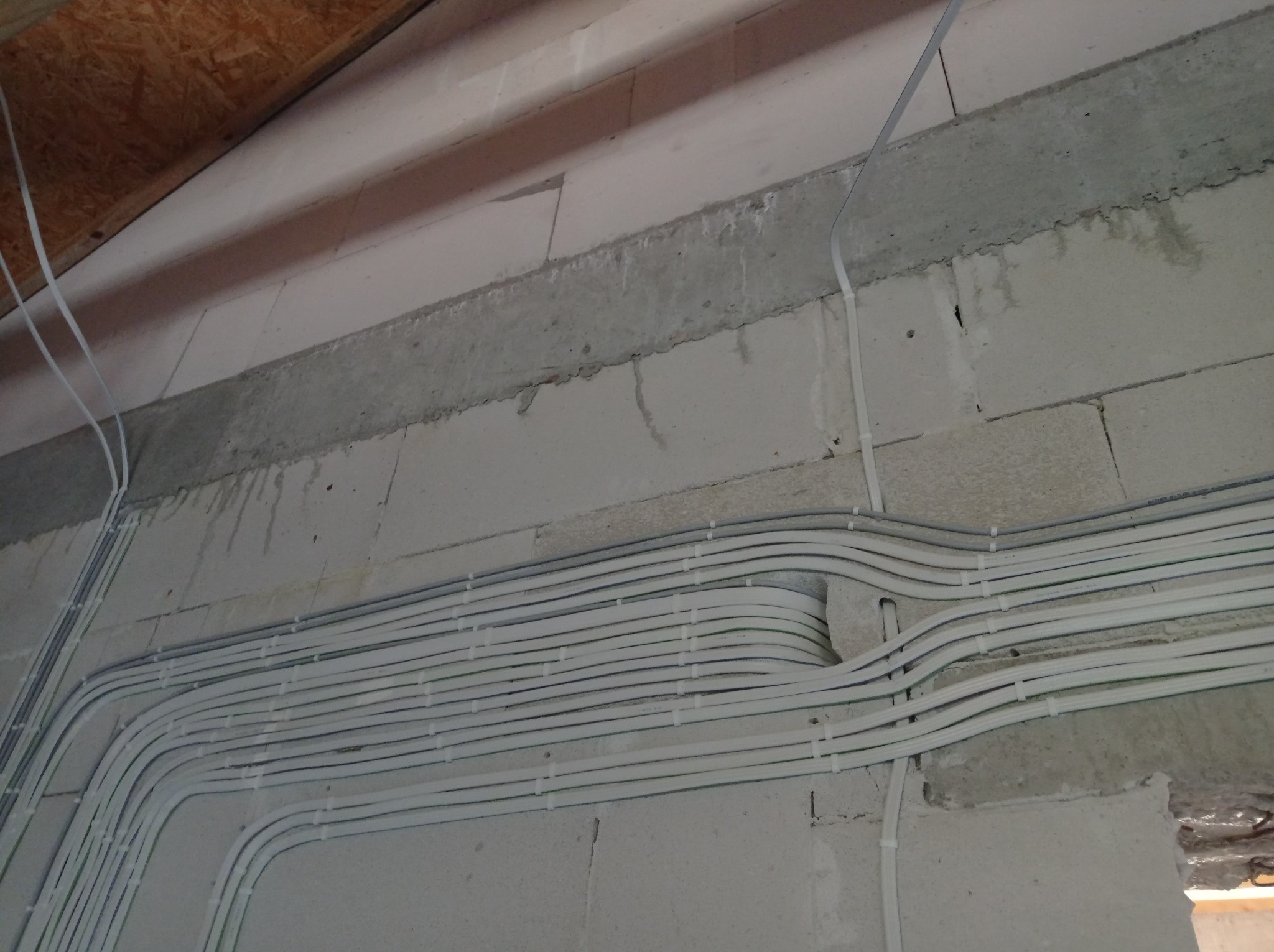 Równolegle ułożone, białe kable elektryczne przymocowane do ściany z betonu komórkowego, widoczne spinki mocujące i fragment drewnianego stropu.