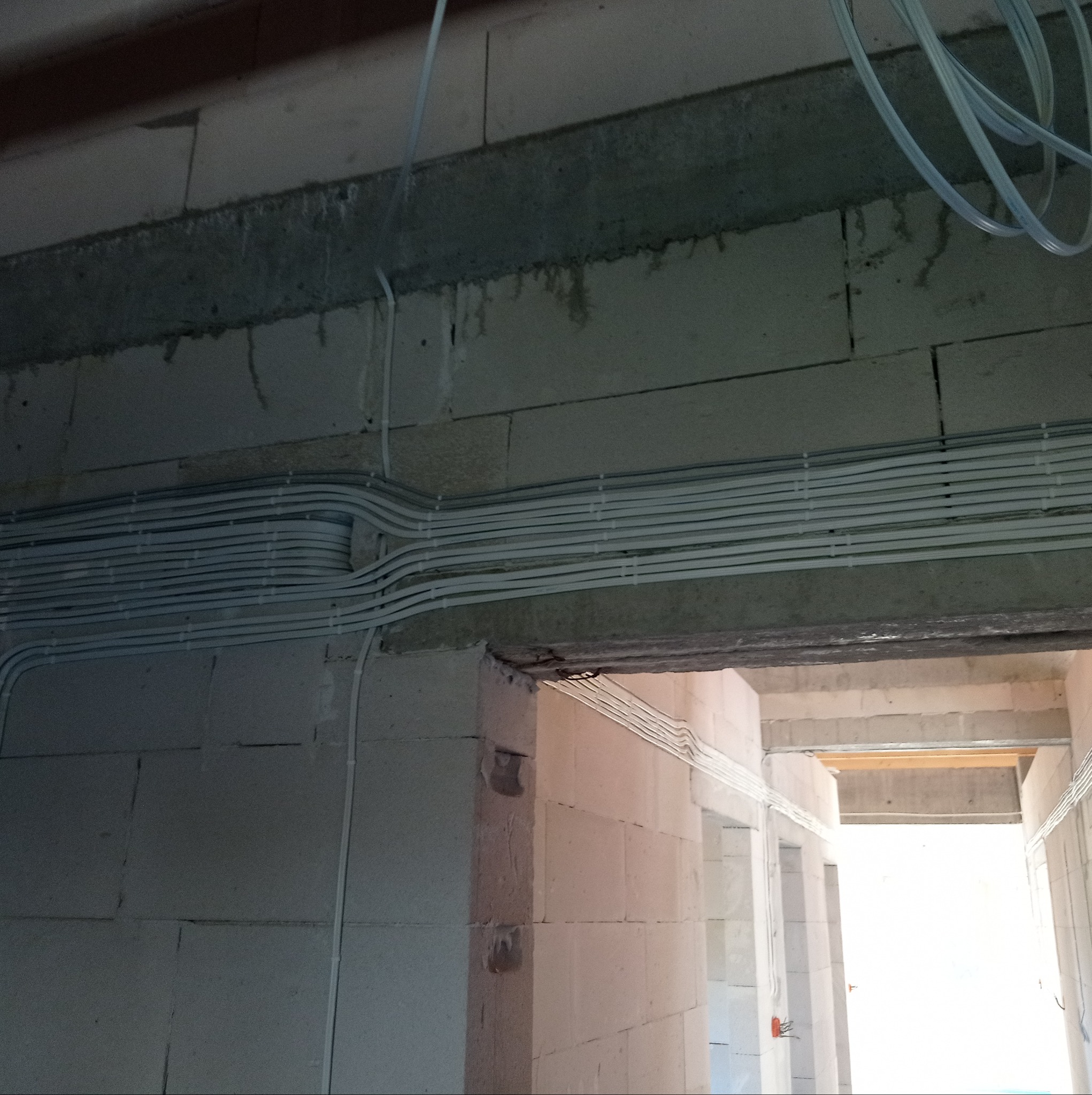 Wiązka przewodów elektrycznych ułożona równolegle pod betonowym stropem w surowym wnętrzu budynku z białych bloczków.