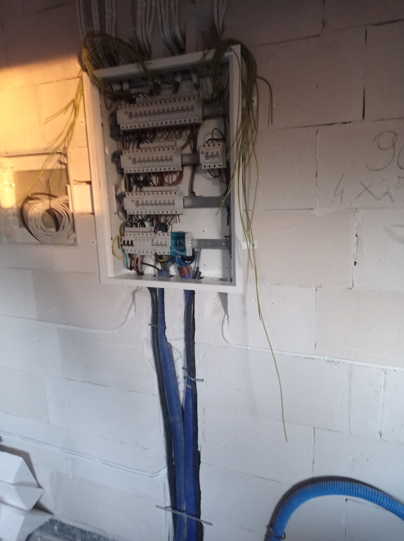 Rozdzielnia elektryczna w trakcie montażu, widoczne kable w peszlach, ściany z betonu komórkowego z notatkami, brakujące elementy obudowy.