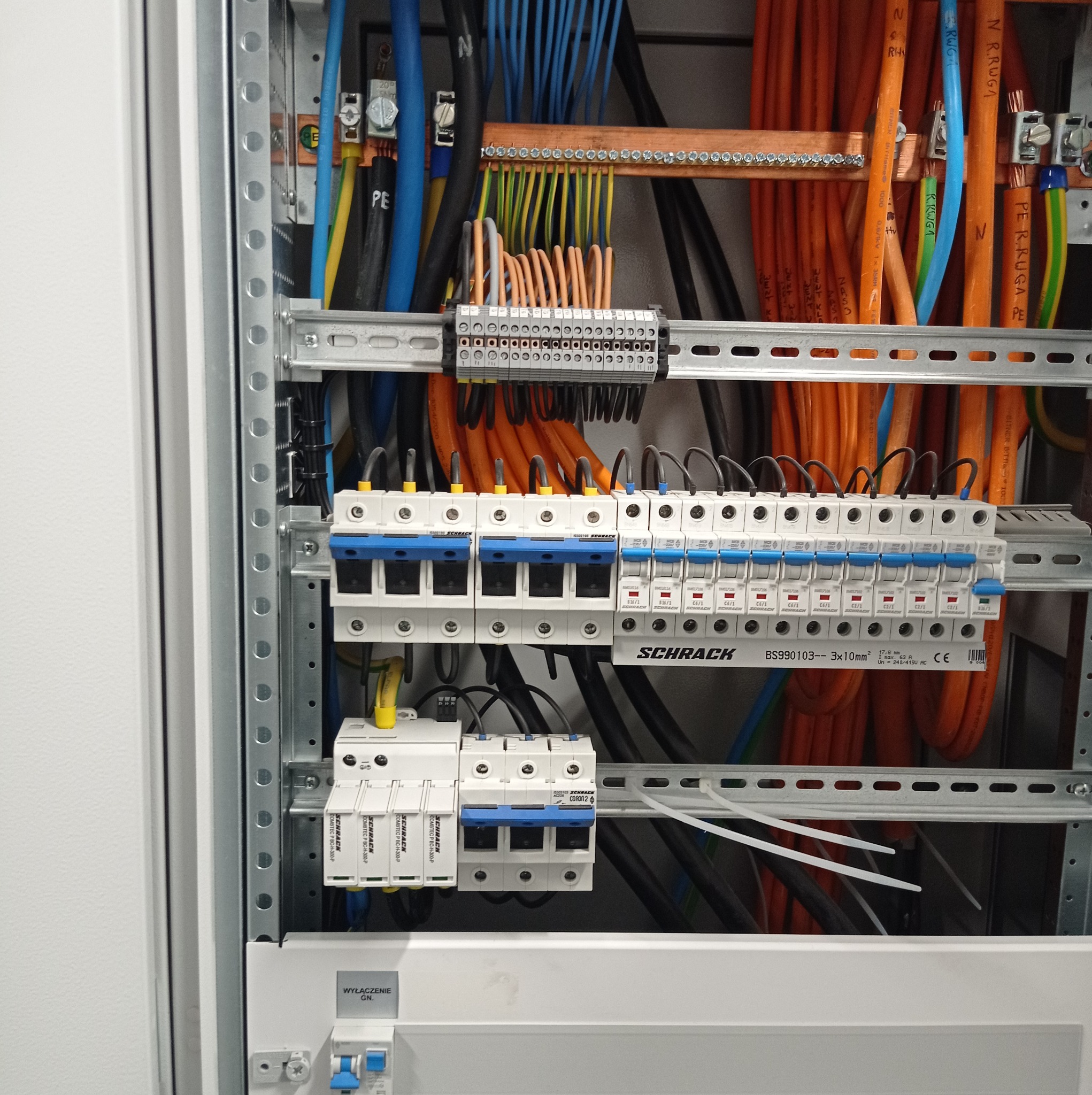 Wnętrze skrzynki elektrycznej z bezpiecznikami Schrack, licznymi przewodami w różnych kolorach (pomarańczowe, niebieskie, czarne, żółto-zielone) i listwami zaciskowymi, zamontowane na szynach DIN.