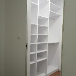 Biała, modułowa garderoba z półkami i drążkami, widoczna z perspektywy wejścia do pomieszczenia; podłoga z paneli imitujących drewno.