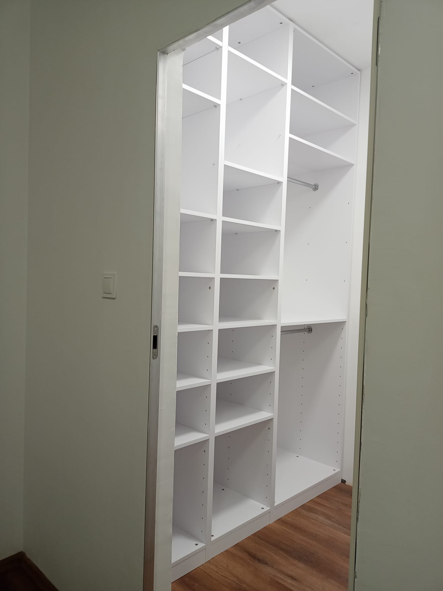 Biała, modułowa garderoba z półkami i drążkami, widoczna z perspektywy wejścia do pomieszczenia; podłoga z paneli imitujących drewno.