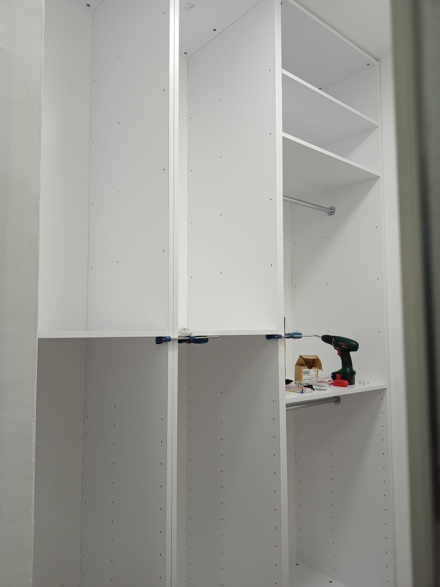 Wnętrze szafy wnękowej w trakcie montażu, widoczne białe płyty, śrubokręt akumulatorowy, pudełko ze śrubami i ściski stolarskie.