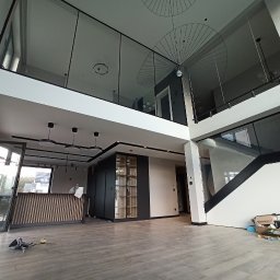 Paweł Grygiel - Wnętrze dwukondygnacyjnego domu w trakcie wykańczania, widoczne przeszklone balustrady na antresoli, schody z czarną poręczą, drewniana podłoga i duże okna, surowe ściany czekające na malowanie.