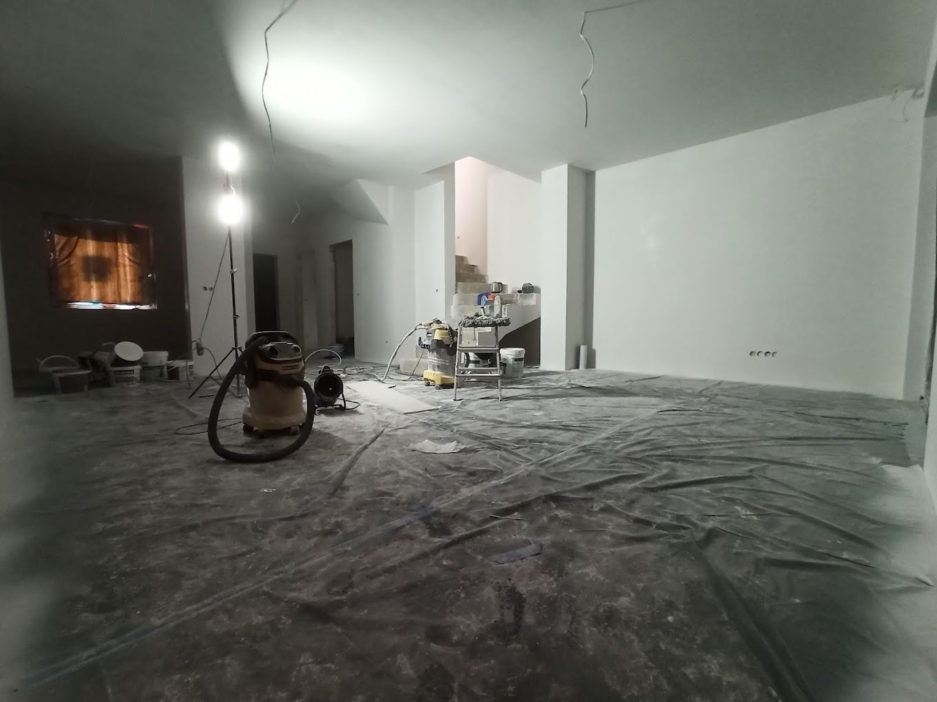 Wnętrze w trakcie prac wykończeniowych: zabezpieczona folią podłoga, sprzęt budowlany, oświetlenie robocze i świeżo wyszpachlowana ściana w tle.
