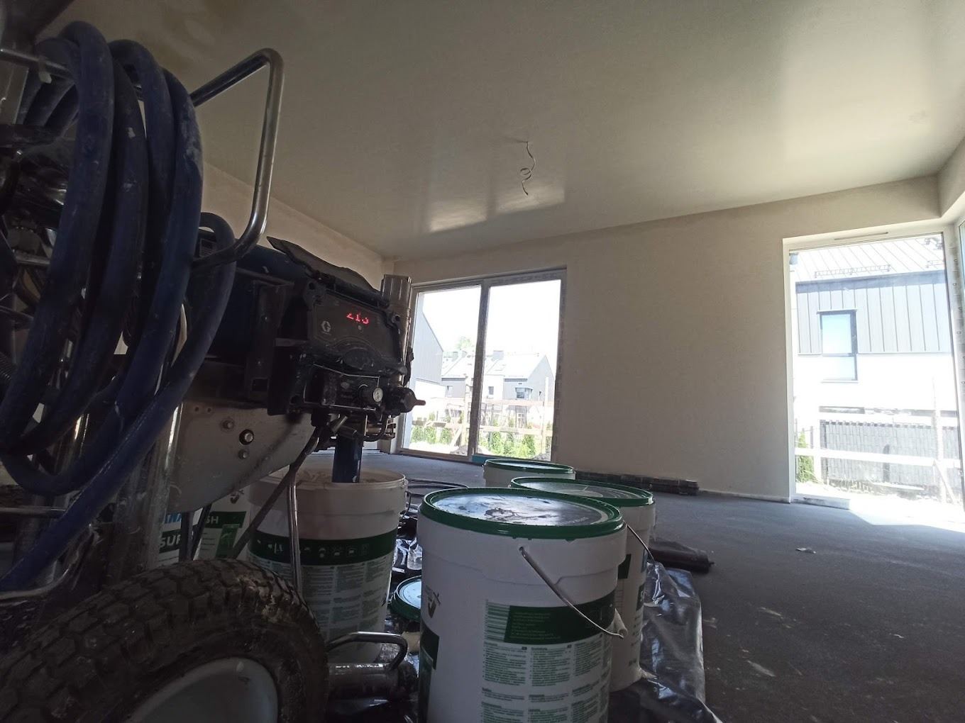 Wnętrze w trakcie prac wykończeniowych: ściany po nałożeniu gładzi, widoczne białe wiadra z farbą i profesjonalny agregat malarski z niebieskim wężem, przygotowane do malowania.