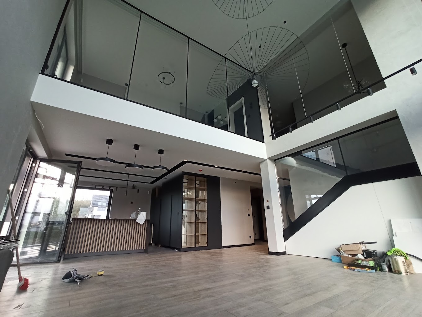 Wnętrze dwukondygnacyjnego domu w trakcie wykańczania, widoczne przeszklone balustrady na antresoli, schody z czarną poręczą, drewniana podłoga i duże okna, surowe ściany czekające na malowanie.