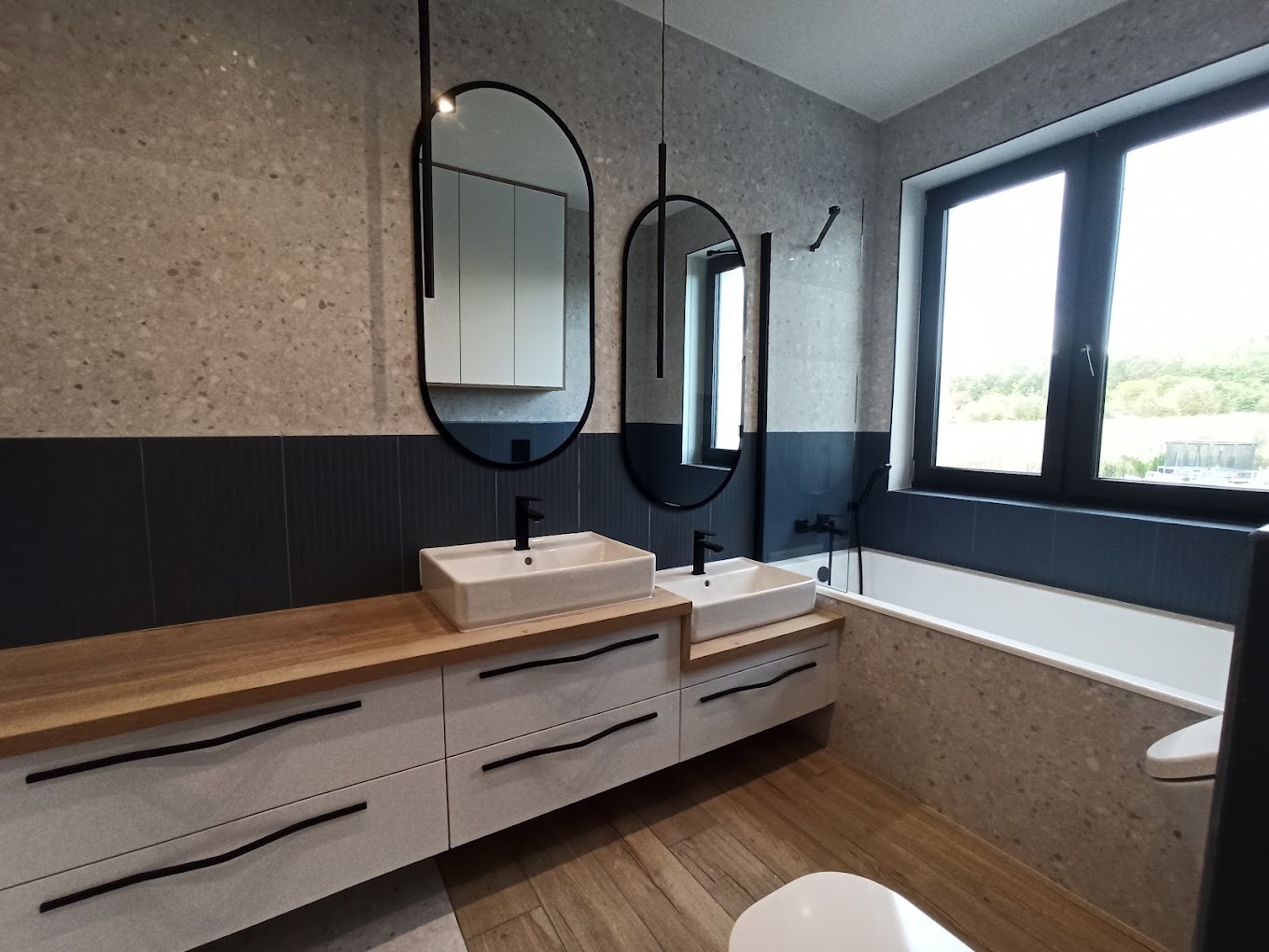 Nowoczesna łazienka z dwoma umywalkami nablatowymi, drewnianym blatem, białymi szafkami z czarnymi uchwytami, dwoma owalnymi lustrami w czarnych ramach i wanną przy oknie.