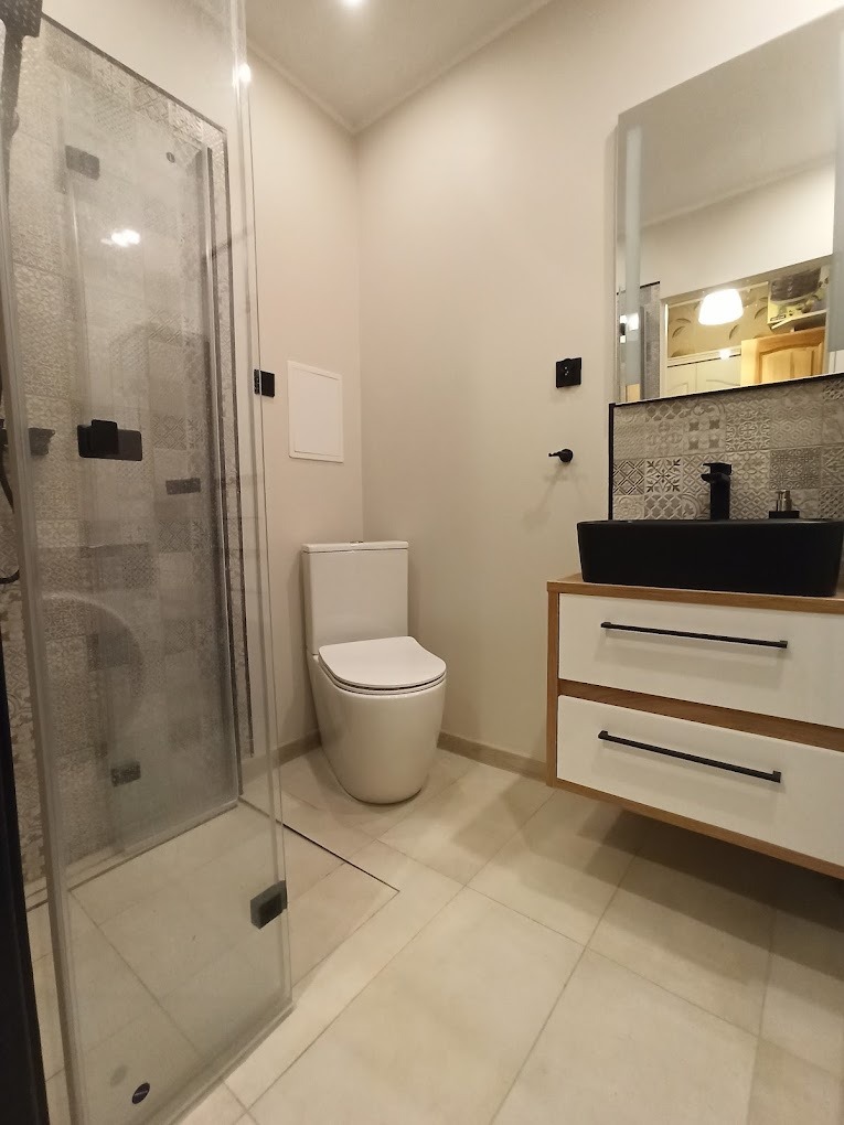 Nowoczesna łazienka z kabiną prysznicową, wiszącą szafką z czarną umywalką i lustrem, oraz toaletą w jasnej kolorystyce.