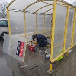 Pracownik w kombinezonie i masce spawalniczej klęczy w żółtej konstrukcji w trakcie spawania, obok leży spawarka Bester i narzędzia, na zewnątrz pada śnieg.