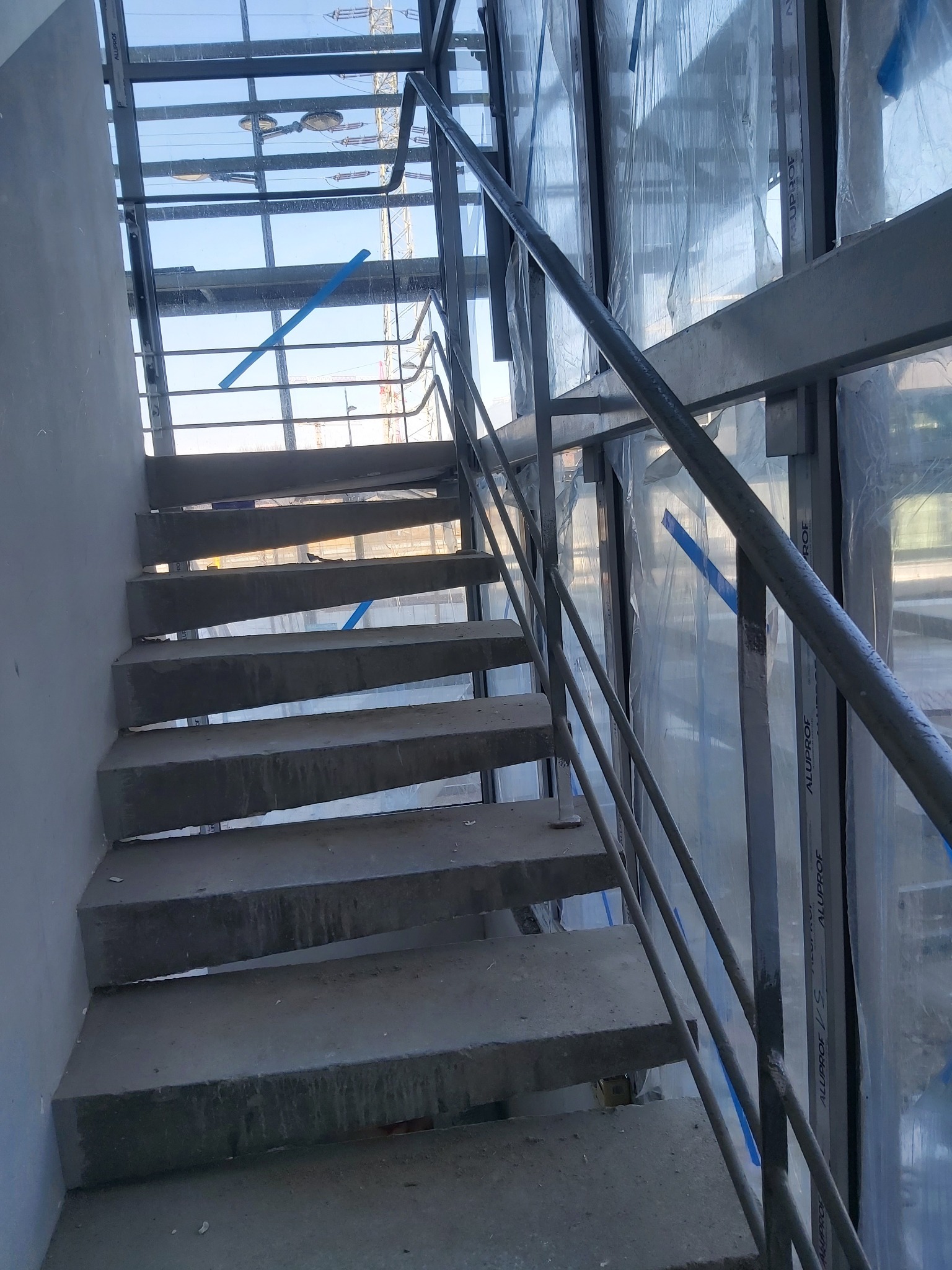 Surowe betonowe schody z metalową balustradą w trakcie budowy, widoczne okna zabezpieczone folią i konstrukcja stalowa dachu.