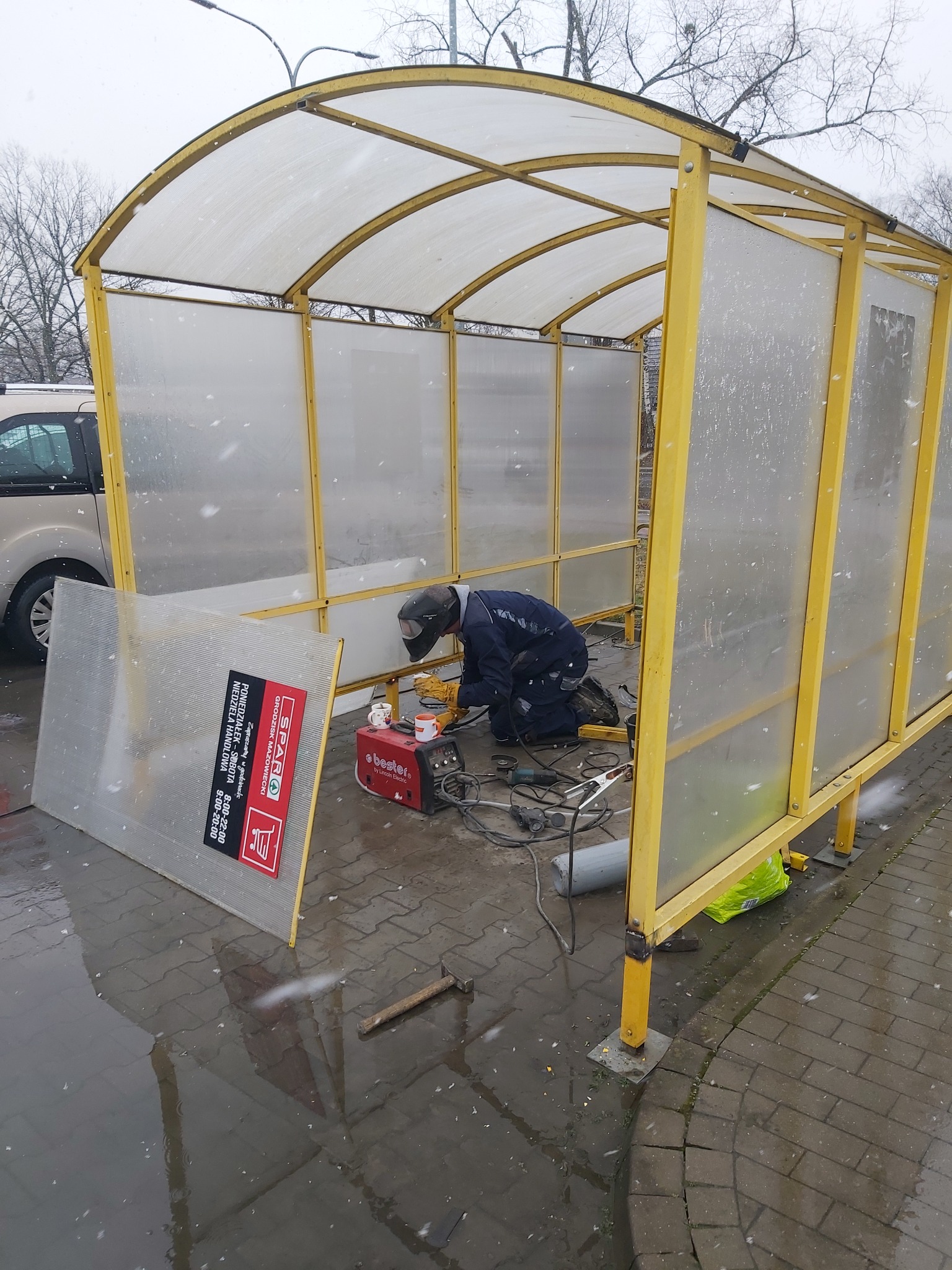 Pracownik w kombinezonie i masce spawalniczej klęczy w żółtej konstrukcji w trakcie spawania, obok leży spawarka Bester i narzędzia, na zewnątrz pada śnieg.