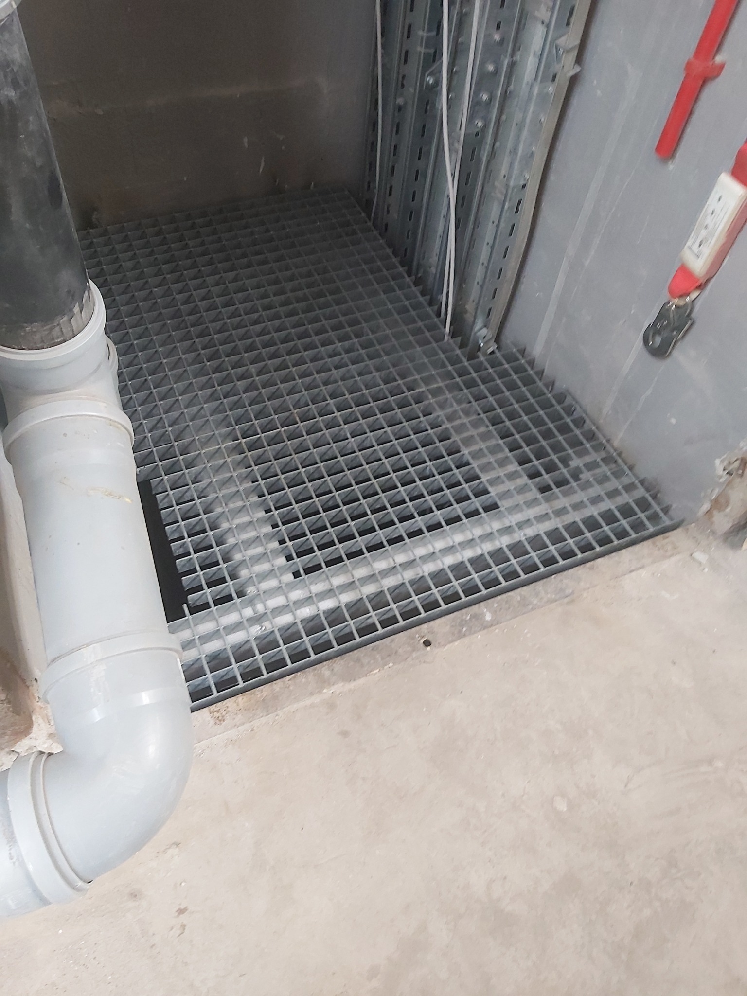 Metalowa krata podłogowa w przestrzeni technicznej, z widocznymi rurami PCV i elementami instalacji elektrycznej na ścianie.