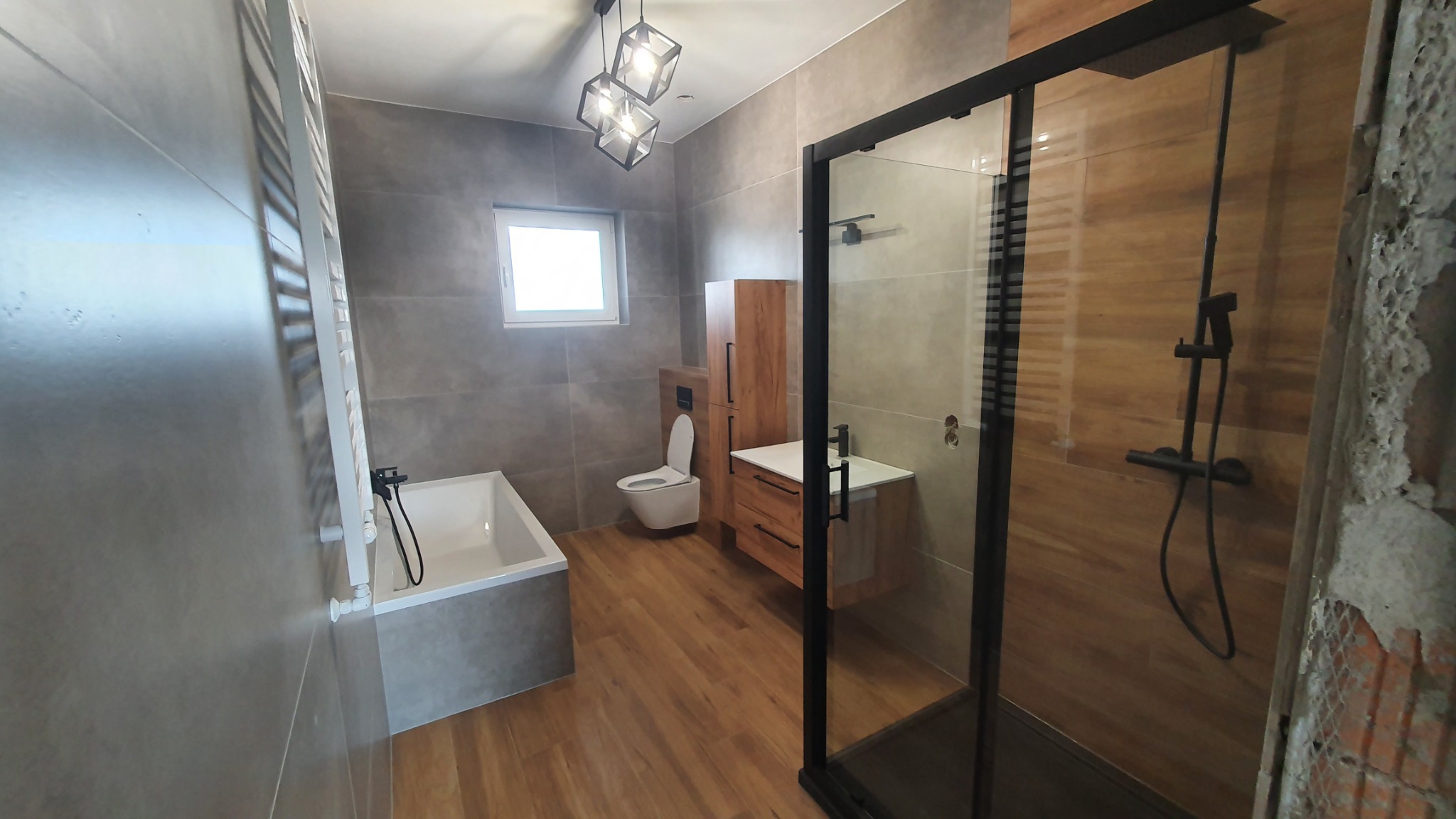 Nowoczesna łazienka po remoncie w Pleszewie, z kabiną prysznicową typu walk-in z czarnymi ramami, drewnopodobnymi płytkami na ścianie prysznica oraz szarymi płytkami na pozostałych ścianach...