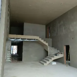 Wnętrze w stanie surowym: ściany pokryte świeżym tynkiem, betonowe schody, widoczne narzędzia i drabina, surowe oświetlenie.