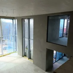 Wnętrze w stanie deweloperskim z surowymi ścianami po nałożeniu tynku, widoczne okna z folią ochronną i wystające kable z sufitu.