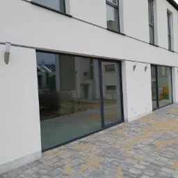 Nowoczesny budynek z białym tynkiem, dużymi oknami z czarnymi ramami i szarą kostką brukową na zewnątrz, z zamontowanymi lampami elewacyjnymi w kształcie tuby.