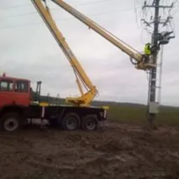 Czerwony, starszy samochód z żółtym podnośnikiem koszowym na błotnistym terenie. Pracownik w kasku i kamizelce na wysokości słupa energetycznego, wykonuje prace konserwacyjne.