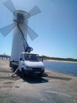 Podnośnik koszowy na podwoziu samochodu dostawczego podczas prac konserwacyjnych przy zabytkowym, białym wiatraku na tle błękitnego nieba i morza.