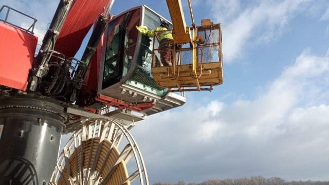 Czerwono-czarny żuraw z żółtą kabiną i koszem, operator w żółtej kamizelce wewnątrz kosza, na tle pochmurnego nieba.