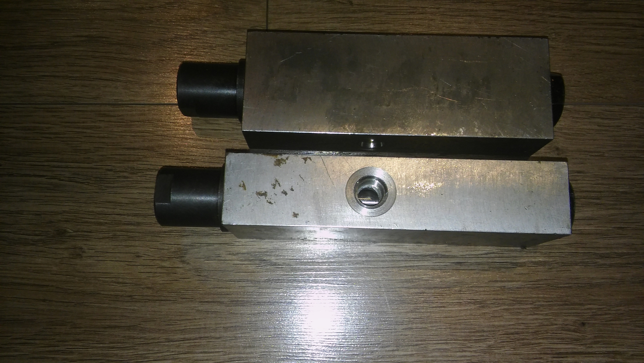Dwa metalowe bloki hydrauliczne leżące na drewnianej powierzchni, widoczne ślady użytkowania i otwór gwintowany w jednym z bloków.
