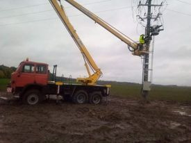 Czerwony, starszy samochód z żółtym podnośnikiem koszowym na błotnistym terenie. Pracownik w kasku i kamizelce na wysokości słupa energetycznego, wykonuje prace konserwacyjne.