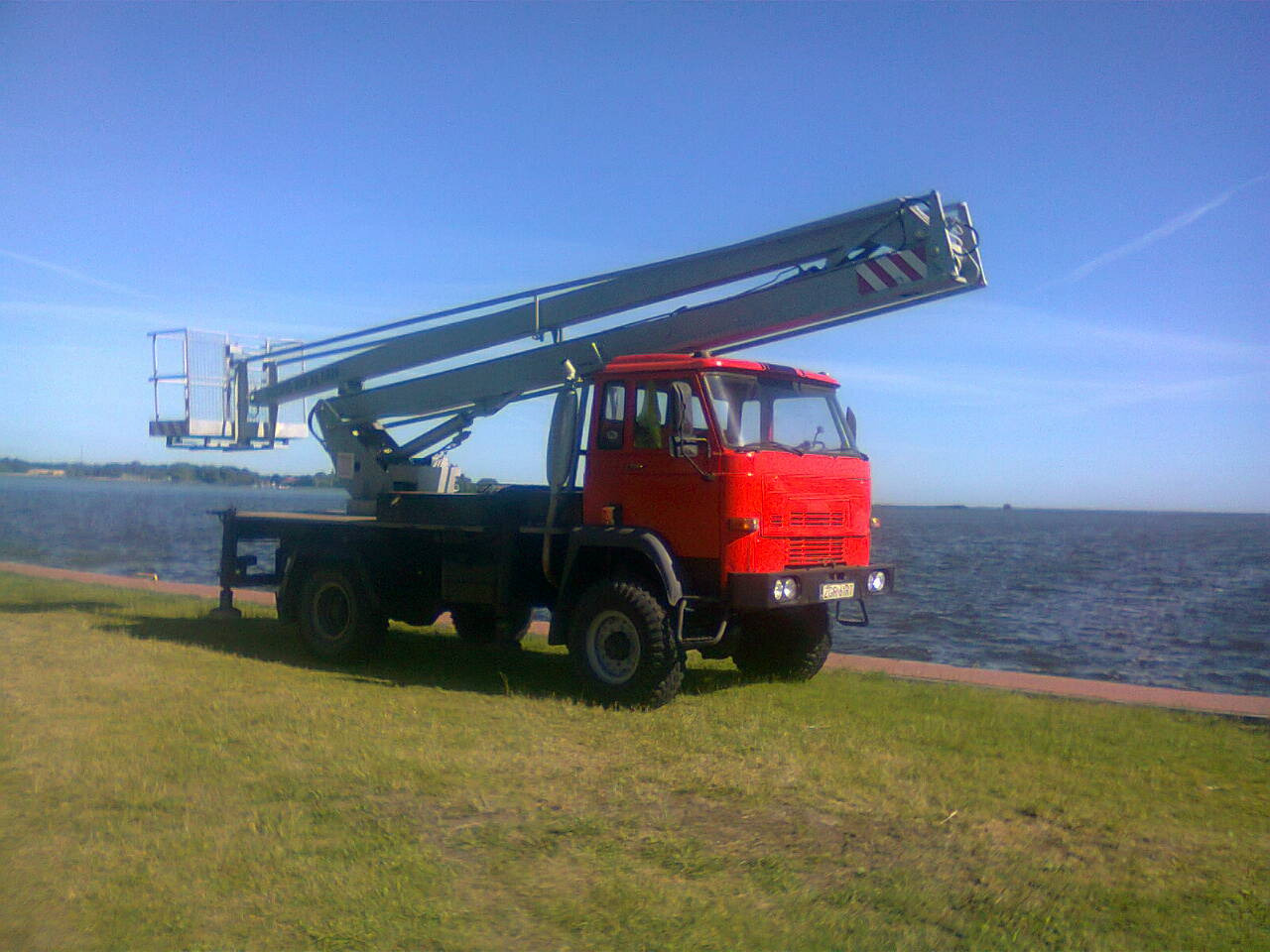 Czerwony samochód ciężarowy z wysięgnikiem koszowym na trawiastym brzegu jeziora w słoneczny dzień.