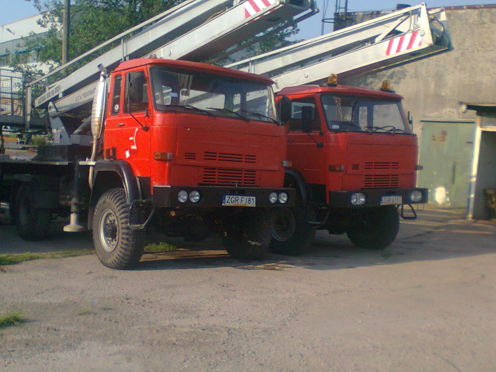 Dwa czerwone samochody ciężarowe z podnośnikami koszowymi zaparkowane na szarym asfalcie, widoczne tablice rejestracyjne ZGR FJ81, w tle budynek z zielonymi drzwiami.