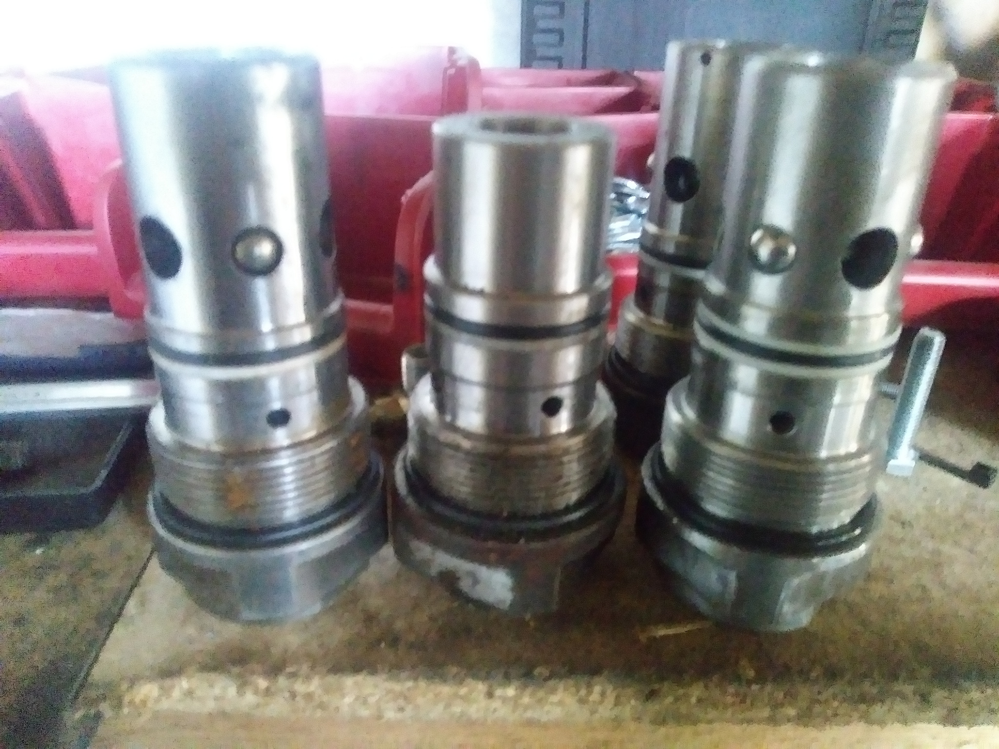 Cztery metalowe, cylindryczne elementy hydrauliczne z otworami i gwintem, prawdopodobnie części zamienne do maszyn budowlanych, ułożone na brudnej powierzchni.