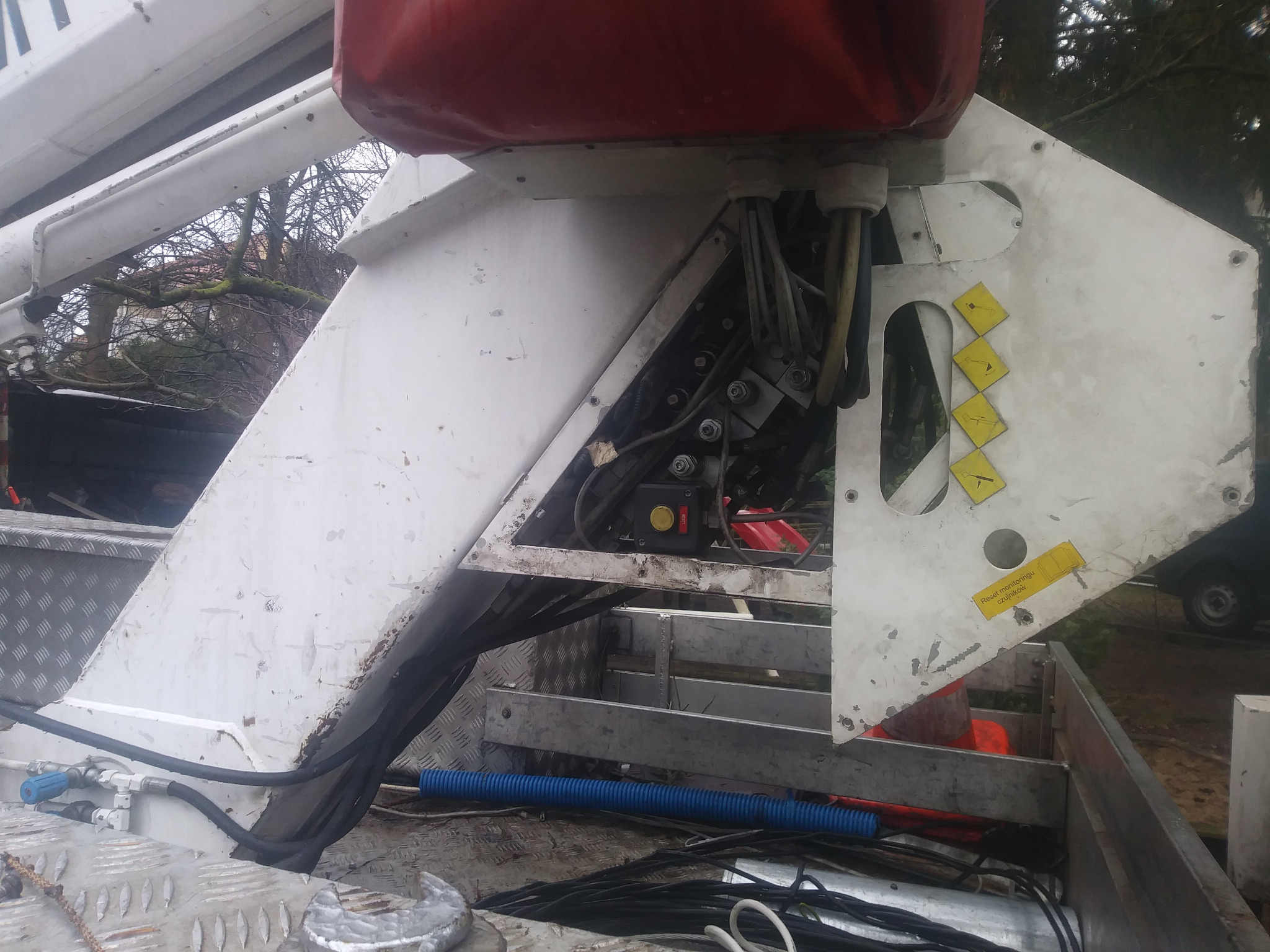 Widok z góry na mechanizm podnośnika koszowego z widocznymi przewodami hydraulicznymi, elementami konstrukcyjnymi i naklejkami ostrzegawczymi, umieszczonego na platformie pojazdu.