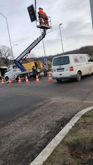Pracownicy w pomarańczowych kombinezonach na podnośniku koszowym naprawiają sygnalizację świetlną na skrzyżowaniu, zabezpieczonym stożkami i pojazdami, w tle drzewa i pochmurne niebo.