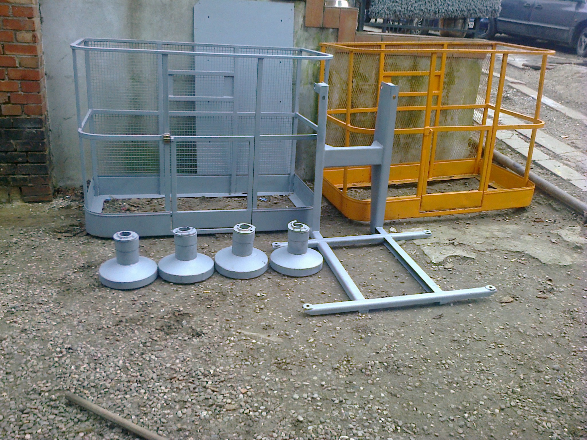 Dwie kosze podnośnikowe, szara i żółta, oraz elementy konstrukcyjne leżą na żwirowym podłożu, gotowe do montażu.