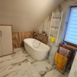 Nowoczesna łazienka na poddaszu z wolnostojącą białą wanną, marmurową podłogą i ścianami, drewnianymi panelami dekoracyjnymi oraz grzejnikiem drabinkowym.