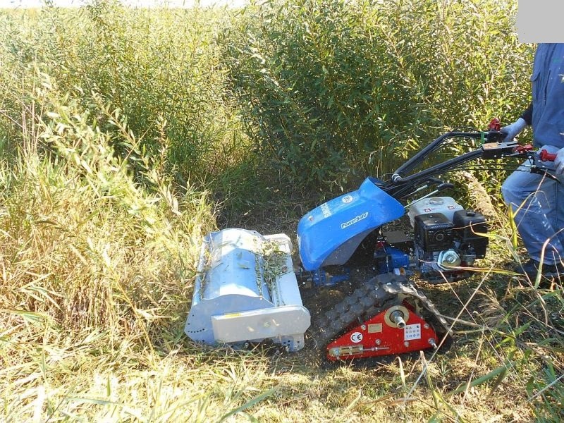 Profesjonalna kosiarka listwowa na gąsienicach w trakcie pracy na zarośniętej łące, widoczne ścięte źdźbła wewnątrz osłony tnącej.