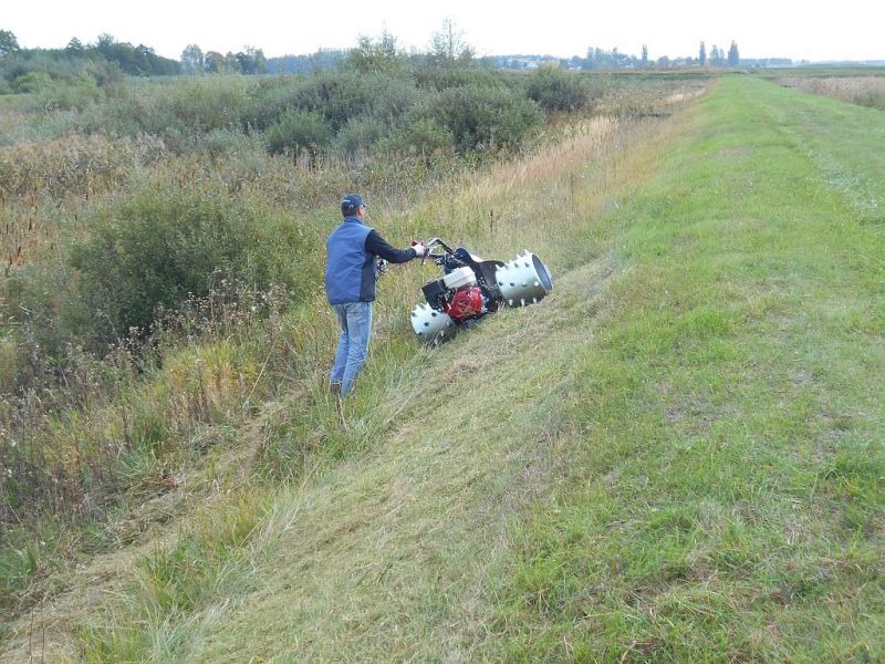 Pracownik w niebieskiej kamizelce obsługuje specjalistyczną kosiarkę bijakową na zboczu rowu melioracyjnego, widoczne świeżo skoszone źdźbła trawy.