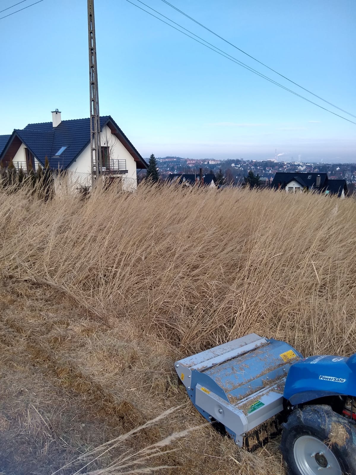 Nisko zawieszony widok na koszenie przerośniętej trawy w Skawinie za pomocą traktorka z kosiarką listwową; w tle domy i zabudowa miejska.
