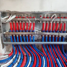 Rozdzielacz ogrzewania podłogowego z rurami w kolorze czerwonym i niebieskim, zamontowany w surowym stanie wnęki ściennej, widoczne pętle rur na powierzchni maty grzewczej.