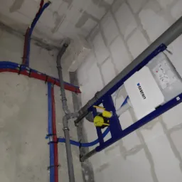 Stelaż podtynkowy Geberit zamontowany w surowym wnętrzu, widoczne rury wodne w kolorze czerwonym i niebieskim oraz szara rura kanalizacyjna.