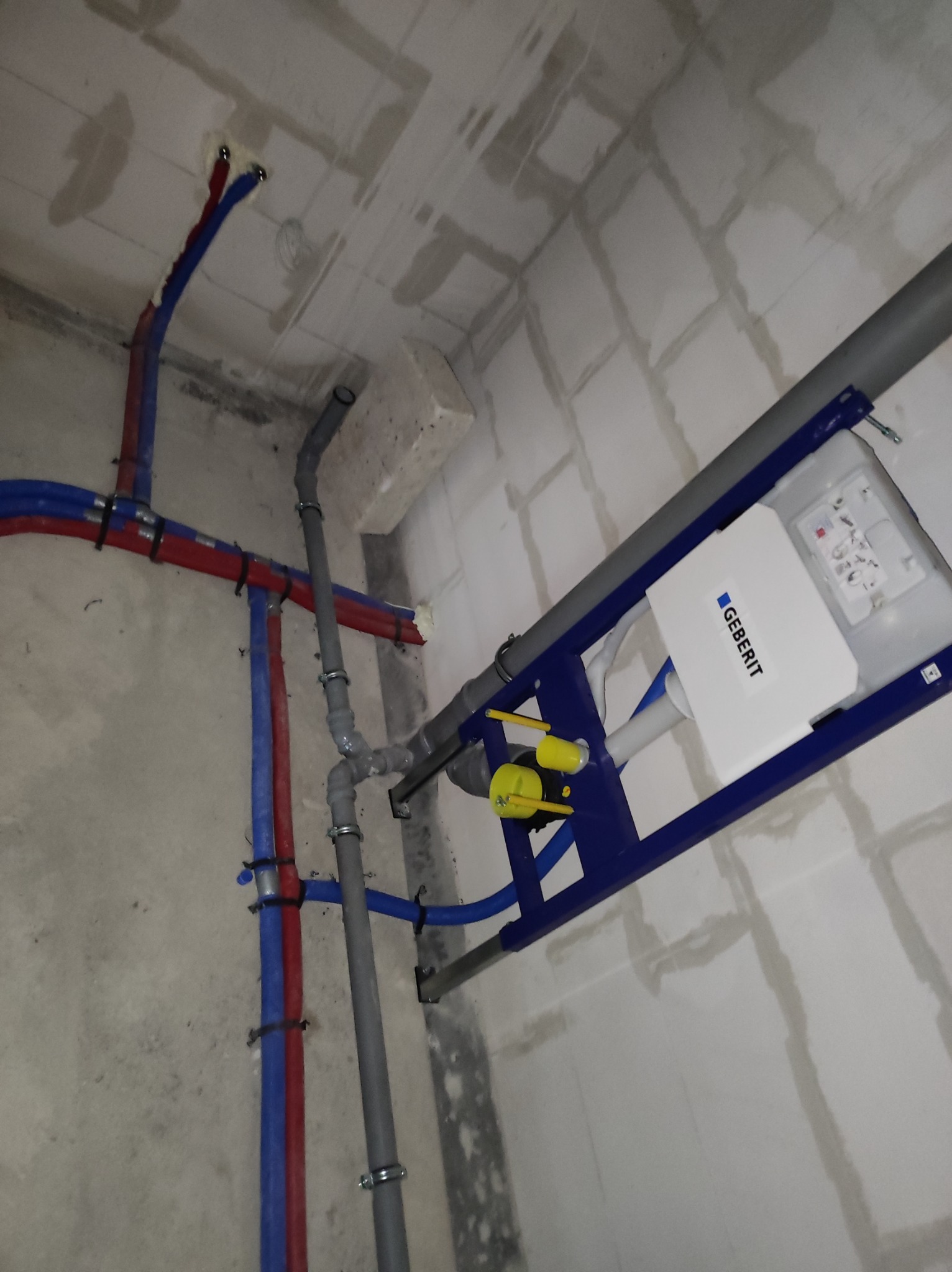 Stelaż podtynkowy Geberit zamontowany w surowym wnętrzu, widoczne rury wodne w kolorze czerwonym i niebieskim oraz szara rura kanalizacyjna.